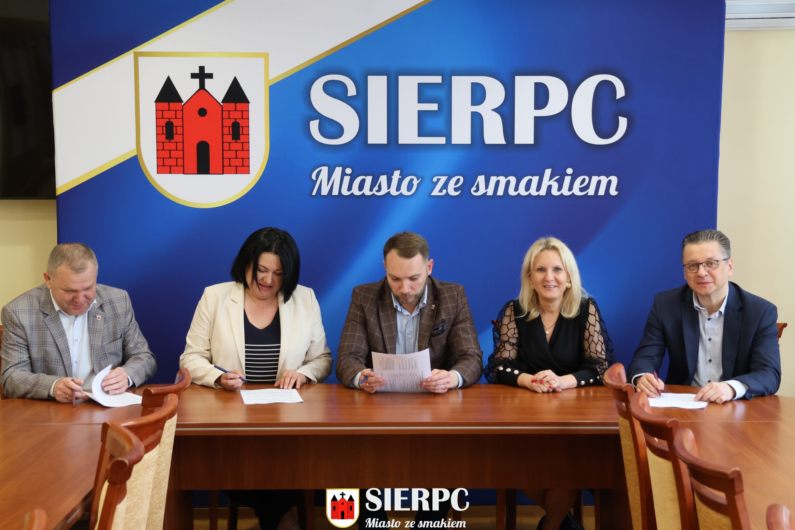 Na zdjęciu widocznych jest 5 osób podpisujących umowę. W tle ścianka reklamowa.