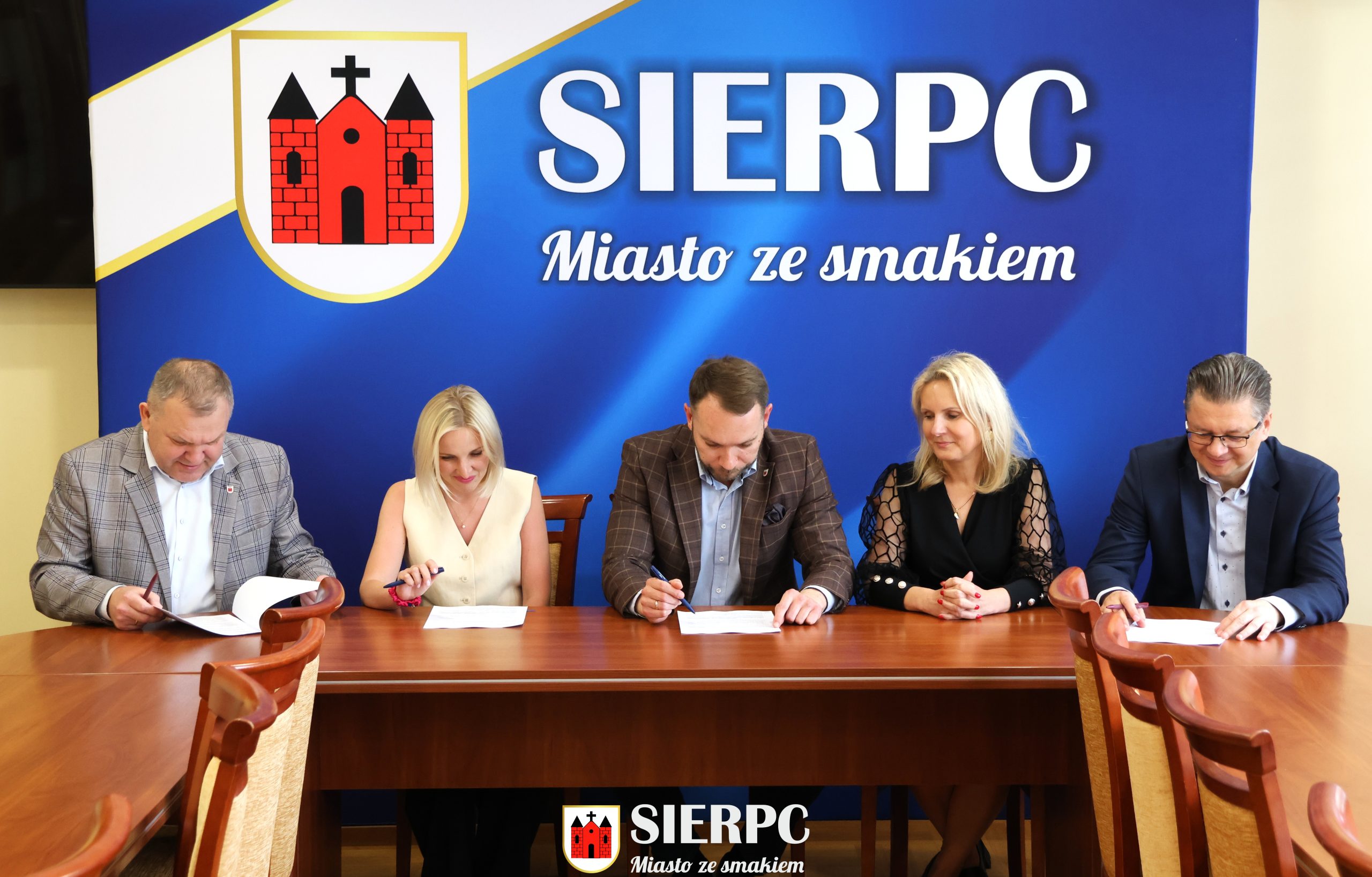 Na zdjęciu widocznych jest 5 osób podpisujących umowę. W tle ścianka reklamowa.