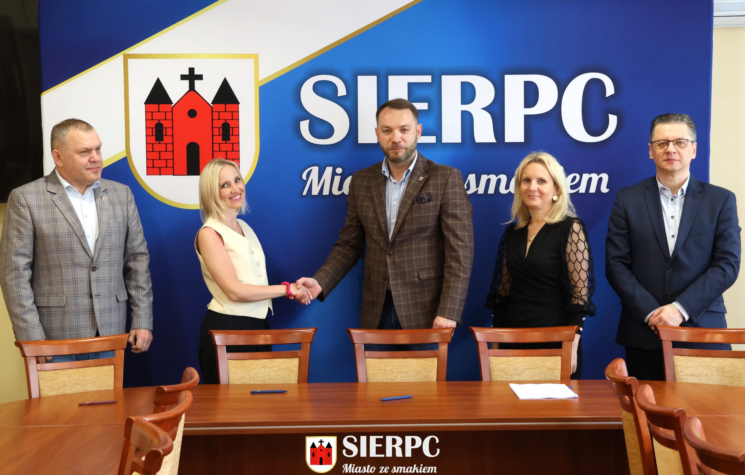 Na zdjęciu widocznych jest 5 osób podpisujących umowę. W tle ścianka reklamowa.