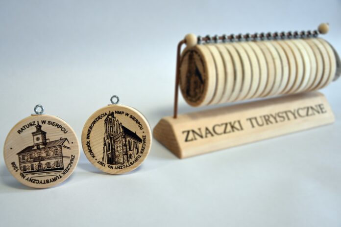 Zdjęcie przedstawia dwa drewniane okrągłe znaczki z wizerunkiem od lewej ratusz w Sierpcu, kolejno Sierpecki Klasztor w tle widoczna większa ilość znaczków zawieszona na drewnianym stojaku
